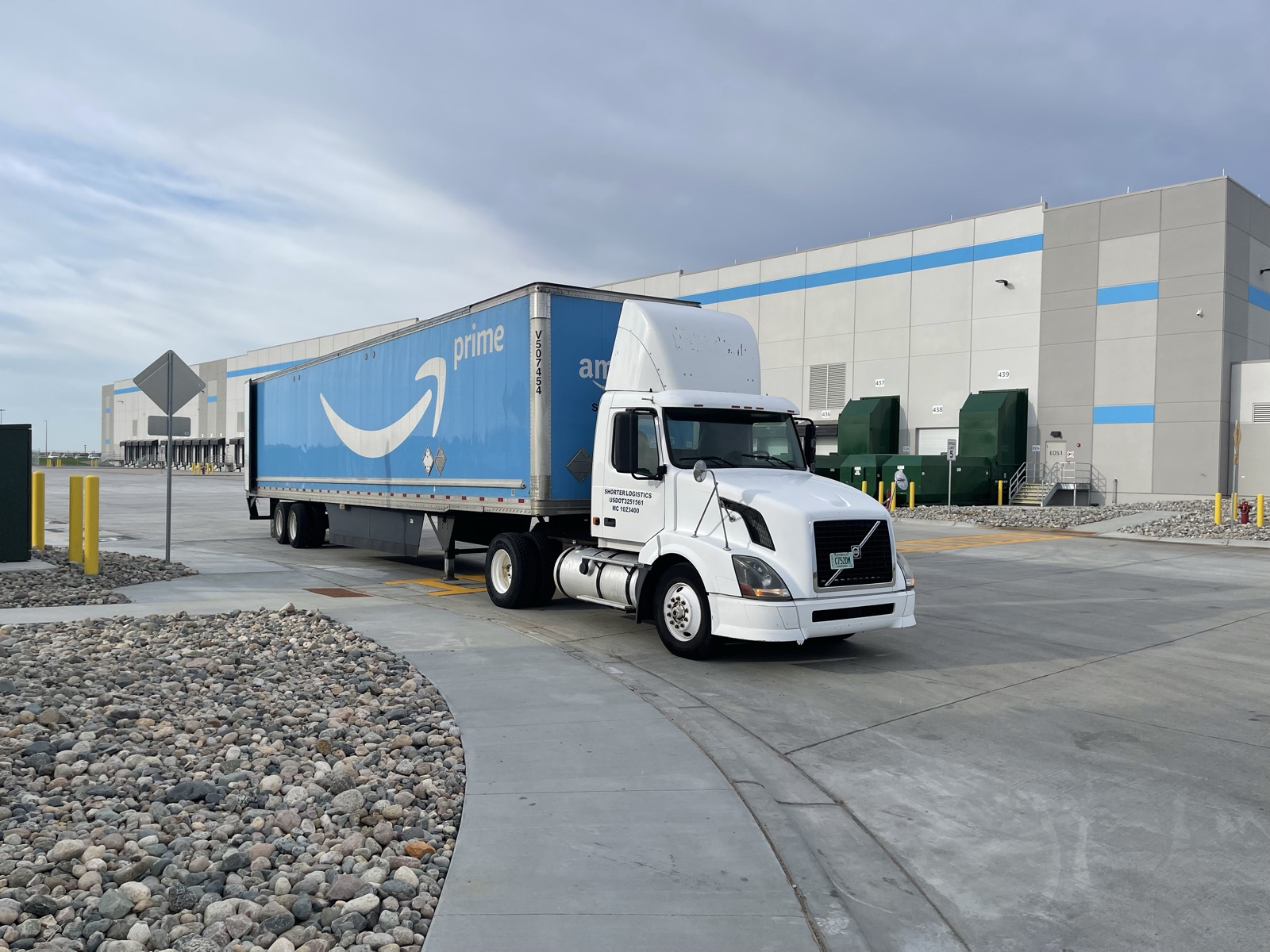 A white Shorter Logistics single-axle day cab pulling a 53-foot trailer past the dock doors of a fulfillment center.